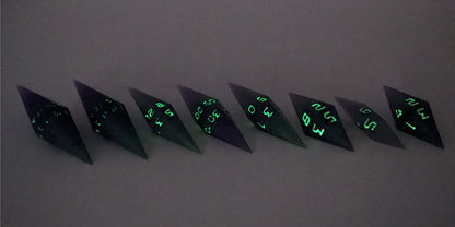A mid angle photo of a complete set of handmade diamond DnD dice that are purple and green with black numbers that glow in the dark arranged in a straight line with bottom points angled slightly forwards and towards the right. They are lined up with the 2 d20s on the left going down to the d4 on the right side. The highest value number of each die is facing forward. The photo is taken in the dark to show the black numbers glowing green.