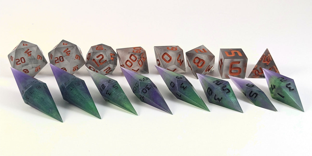 A straight on photo of a complete set of dagger dice that are purple and green with black numbers that glow in the dark arranged in a straight line with points angled forward and to the right. They are lined up with the 2 d20s on the left going down to the d4 on the right side. Behind the dagger set is a standard set of polyhedral dice to allow for size comparison. The diamond dice are much smaller overall. The highest value number of each die is facing forward.