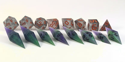 A straight on photo of a complete set of dagger dice that are purple and green with black numbers that glow in the dark arranged in a straight line with points angled forward and to the right. They are lined up with the 2 d20s on the left going down to the d4 on the right side. Behind the dagger set is a standard set of polyhedral dice to allow for size comparison. The diamond dice are much smaller overall. The highest value number of each die is facing forward.