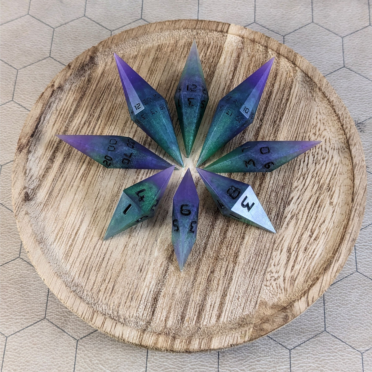 A set of handmade diamond dice that are purple and green with black numbers that glow in the dark arranged on a wooden circle. The resin dice have their tips facing outward, and the highest number of each die is facing up.