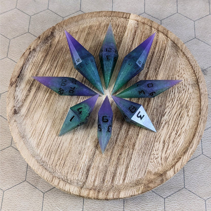 A set of handmade diamond dice that are purple and green with black numbers that glow in the dark arranged on a wooden circle. The resin dice have their tips facing outward, and the highest number of each die is facing up.