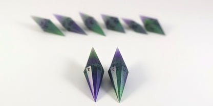 A straight on photo of a diamond set of DnD dice that are purple and green with black numbers that glow in the dark focusing on 2 d20s in the center. The d20 on the left has the 1 face up and the d20 on the right has the 20 face up. The other 6 dice are lined up in the background but are out of focus, while the d20s are in sharp focus.