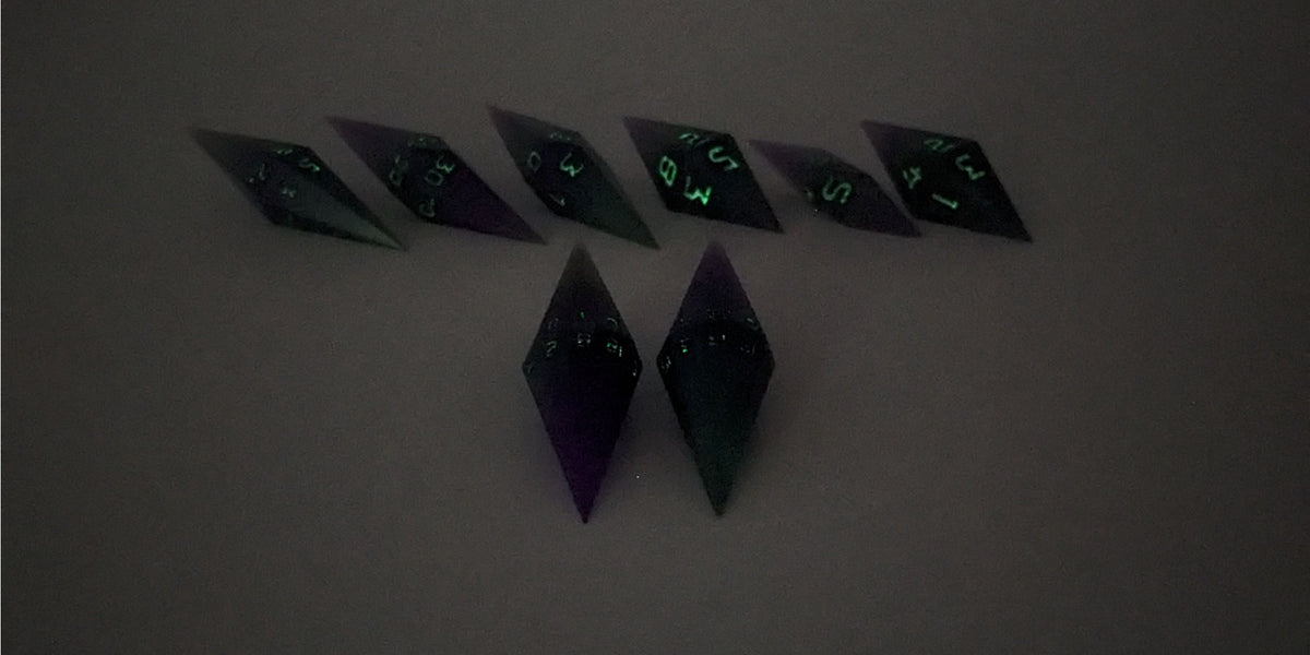 A straight on photo of a diamond set of DnD dice that are purple and green with black numbers that glow in the dark focusing on 2 d20s in the center. The d20 on the left has the 1 face up and the d20 on the right has the 20 face up. The other 6 dice are lined up in the background but are out of focus, while the d20s are in sharp focus. The photo is taken in the dark to show the black numbers glowing green.