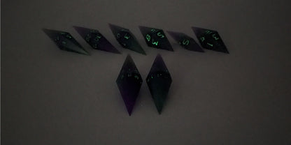 A straight on photo of a diamond set of DnD dice that are purple and green with black numbers that glow in the dark focusing on 2 d20s in the center. The d20 on the left has the 1 face up and the d20 on the right has the 20 face up. The other 6 dice are lined up in the background but are out of focus, while the d20s are in sharp focus. The photo is taken in the dark to show the black numbers glowing green.