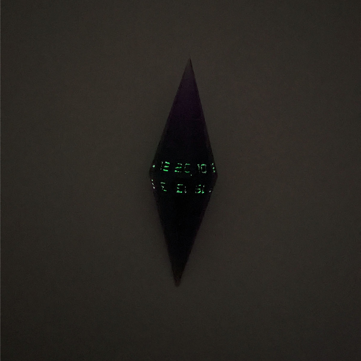 A high angle view of a diamond d20 with swirls of purple and green with black numbers that glow in the dark on a white background. This photo is taken in the dark to show the black numbers glowing green in the dark.