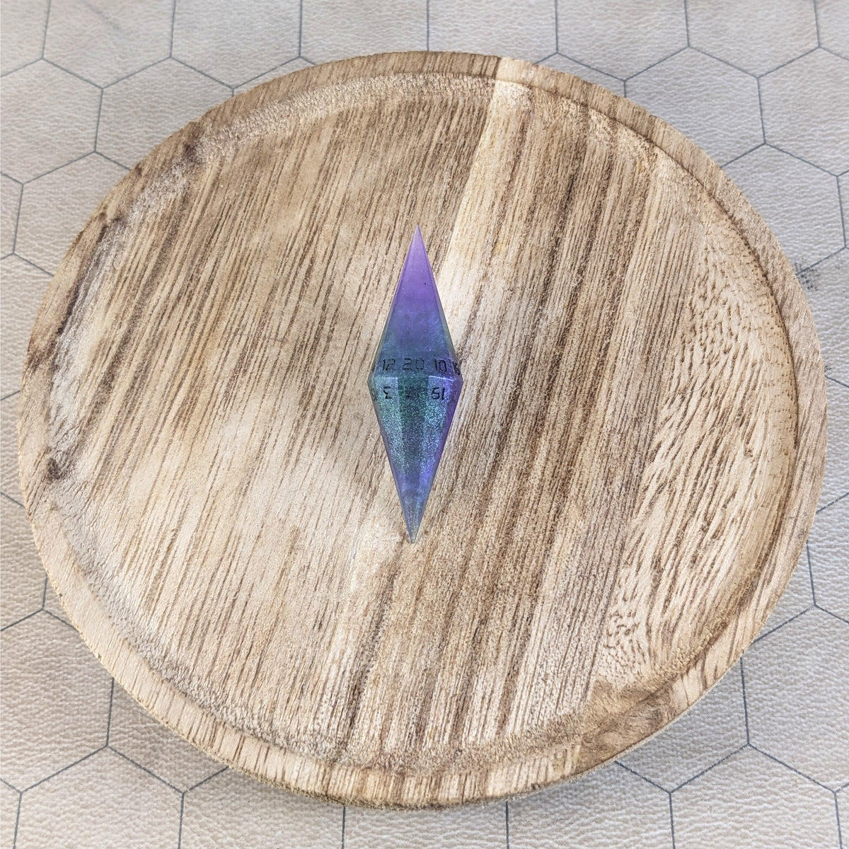 A high angle view of a diamond d20 with swirls of purple and green with black numbers that glow in the dark. It is sitting in the center of a wooden circle.