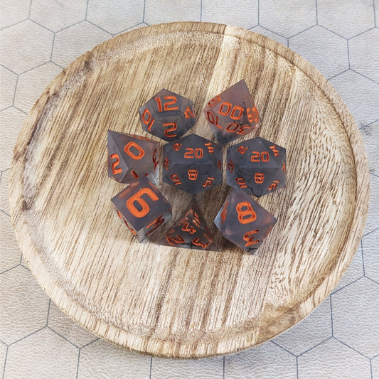 A complete set of 8 handmade artisan dice that have have black swirls with orange numbers. The DnD dice set is on a wooded circle and the dice are arranged in a circle and features 2 D20s, one of which is in the center surrounded by the other 7 dice.