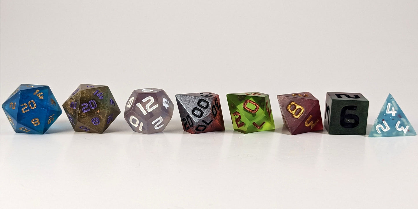 A straight on photo of a complete set of polyhedral dice that are a mismatched array of colors and designs arranged in a straight line. They are lined up with the 2 d20s on the left going down to the d4 on the right side. The highest value number of each die is facing forward.