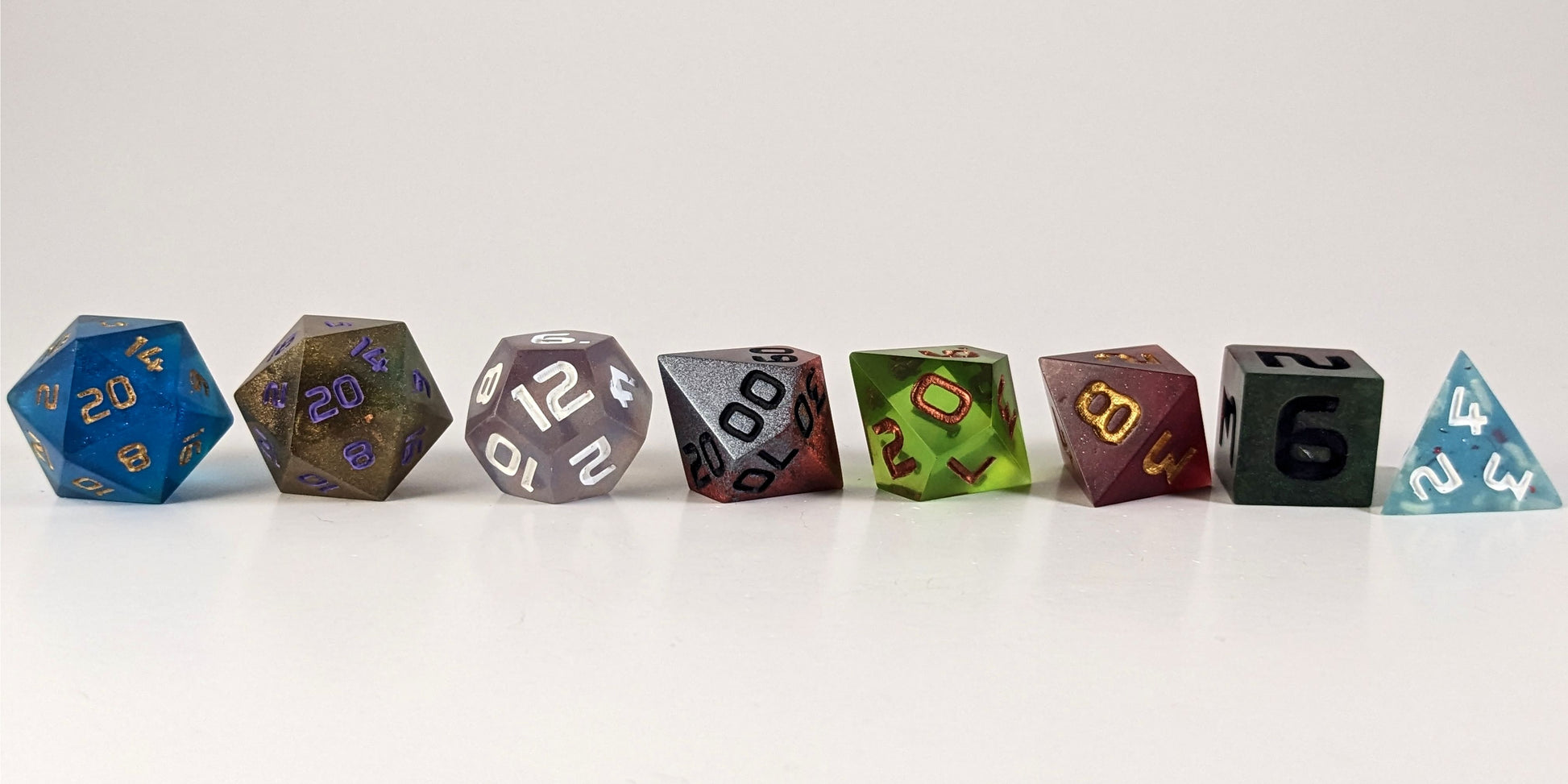 A straight on photo of a complete set of polyhedral dice that are a mismatched array of colors and designs arranged in a straight line. They are lined up with the 2 d20s on the left going down to the d4 on the right side. The highest value number of each die is facing forward.