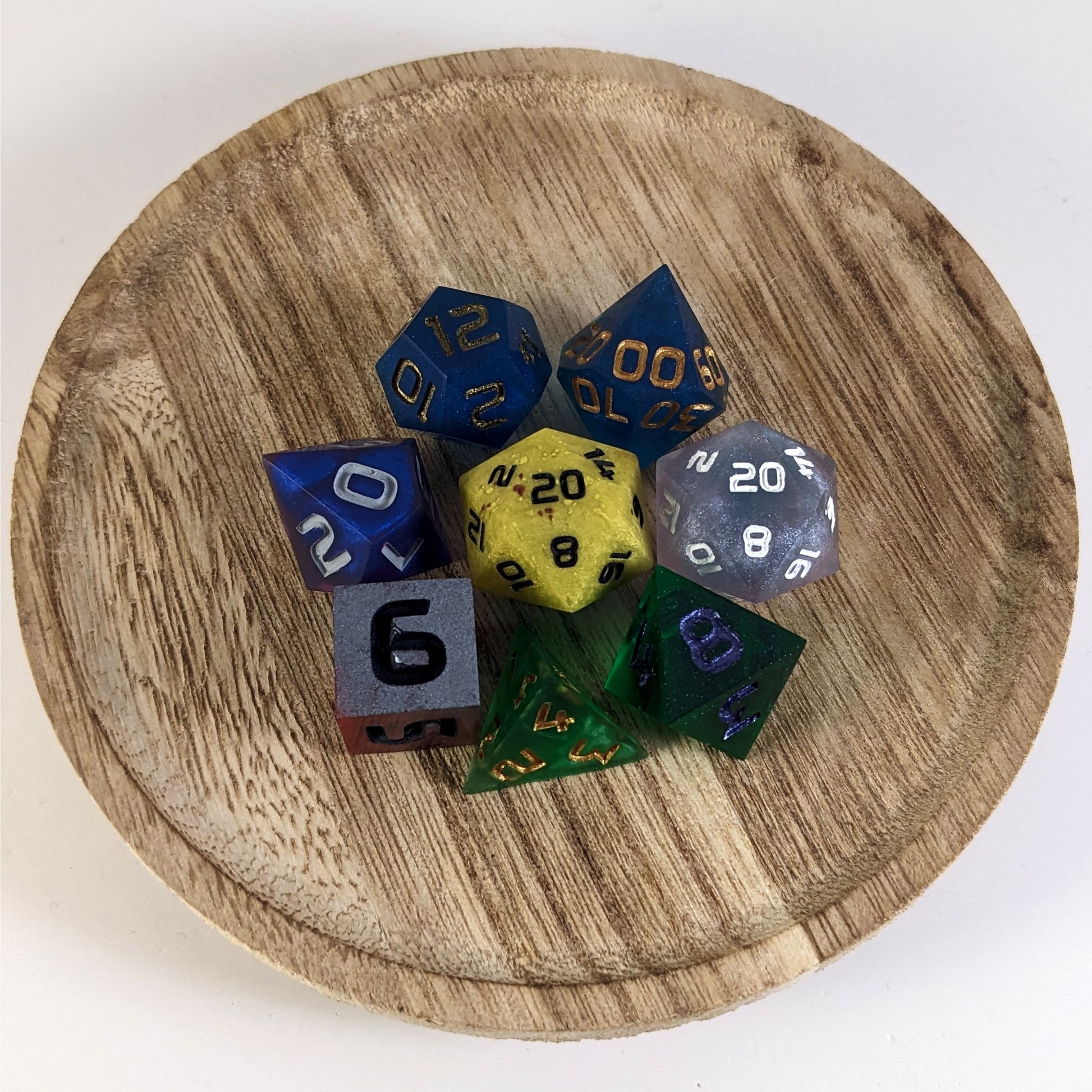 A complete set of 8 handmade artisan dice that are a mismatched array of different colors and designs. The DnD dice set is on a wooden circle and the dice are arranged in a circle and features 2 D20s, one of which is in the center surrounded by the other 7 dice.