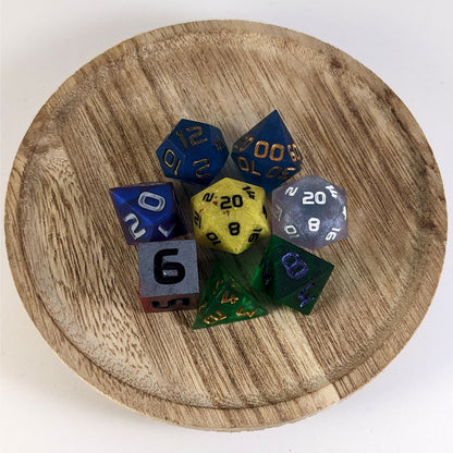 A complete set of 8 handmade artisan dice that are a mismatched array of different colors and designs. The DnD dice set is on a wooden circle and the dice are arranged in a circle and features 2 D20s, one of which is in the center surrounded by the other 7 dice.