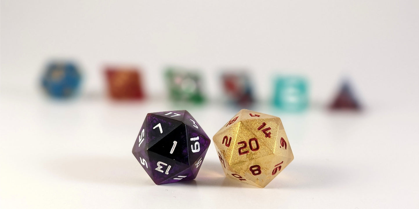 A straight on photo of a standard set of dice that are a mismatched array of colors and designs focusing on 2 d20s in the center. The d20 on the left has the 1 face centered and the d20 on the right has the 20 face centered. The other 6 dice are lined up in the background but are out of focus, while the d20s are in sharp focus.