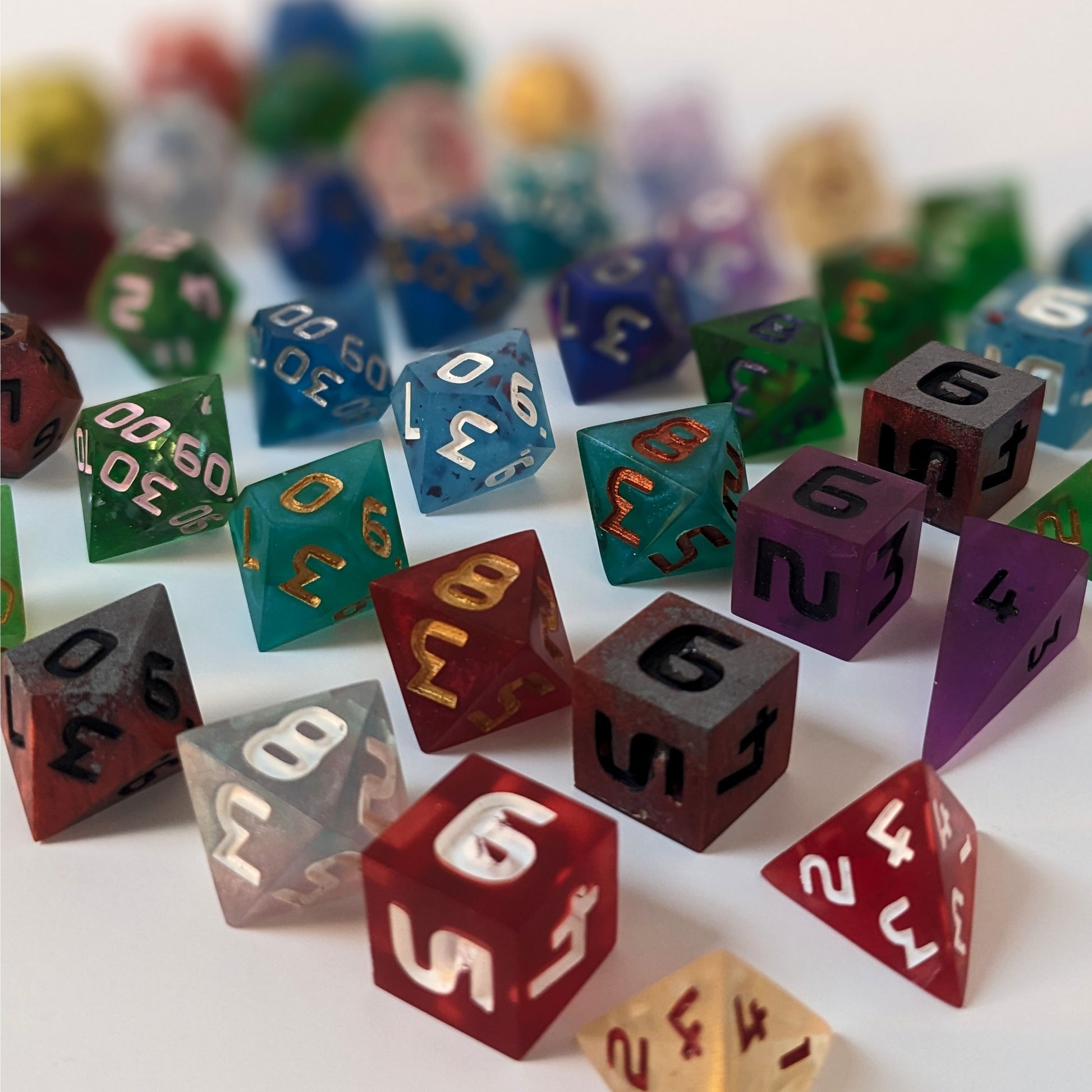 Multiple sets of polyhedral dnd dice lined up in face number order. The dice are a variety of colors and designs and sit on a white background. The photo is taken at an angle with the d4s and d6s closest and angles back so that the d20s and d12s are out of focus in the back of the image.