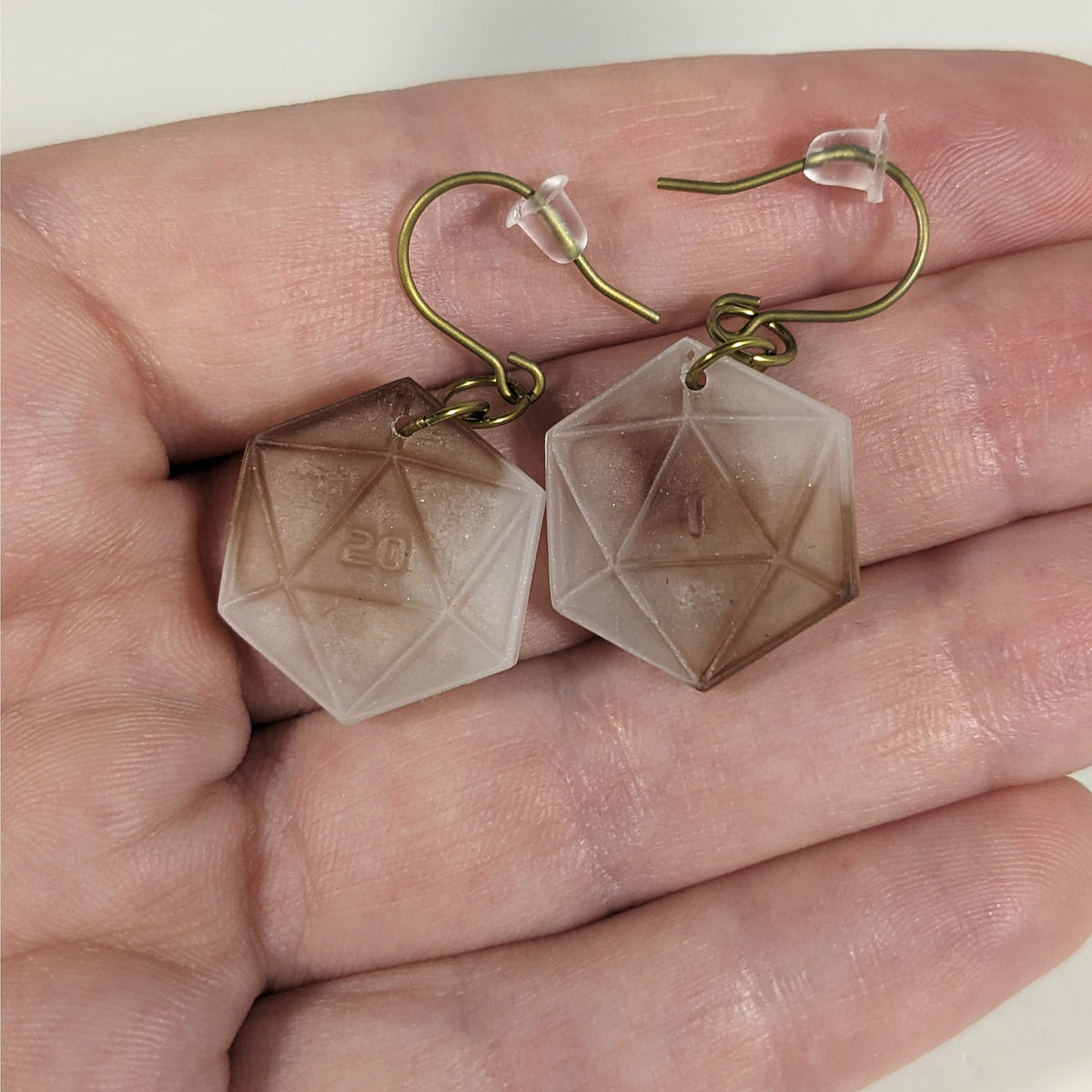A pair of espresso brown and white dangly, flat d20 earrings with gold color titanium and niobium hardware with bell shaped rubber backs held on on a hand on a white background. The earring on the left has a 20 on it and the earring on the right has a 1 on it, both in a sci-fi style font.