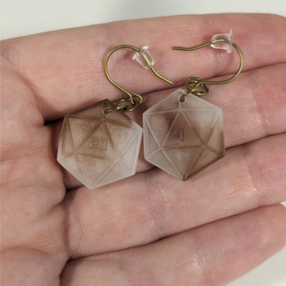 A pair of espresso brown and white dangly, flat d20 earrings with gold color titanium and niobium hardware with bell shaped rubber backs held on on a hand on a white background. The earring on the left has a 20 on it and the earring on the right has a 1 on it, both in a sci-fi style font.