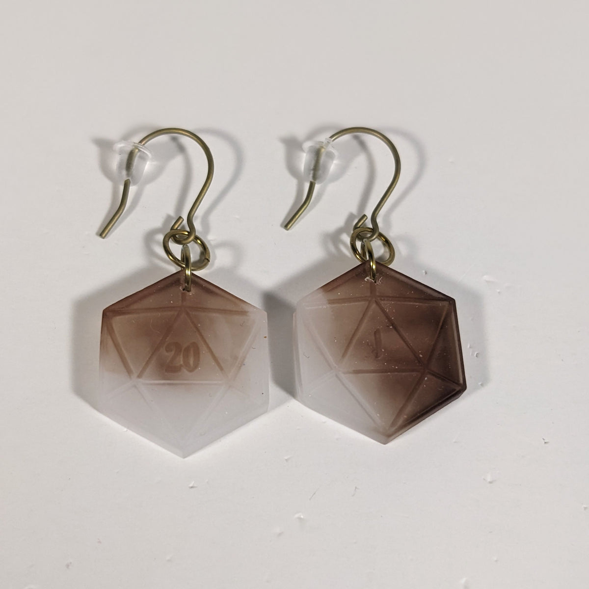 A pair of espresso brown and white dangly, flat d20 earrings with gold color titanium and niobium hardware and bell shaped rubber backs on a white background. The earring on the left has a 20 on it and the earring on the right has a 1 on it, both in a fantasy style font.