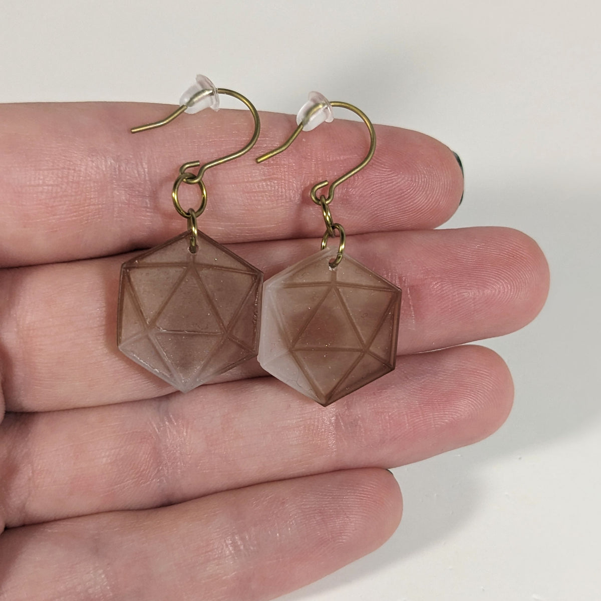 A pair of espresso brown and white dangly, flat d20 earrings with gold color titanium and niobium hardware with bell shaped rubber backs held on on a hand on a white background.