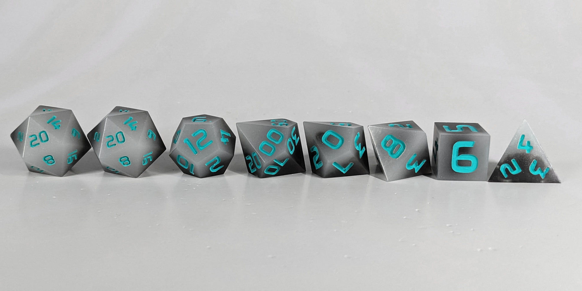A straight on photo of a complete set of polyhedral dice that are coffee brown with milky white swirls and teal numbers arranged in a straight line. They are lined up with the 2 d20s on the left going down to the d4 on the right side. The highest value number of each die is facing forward