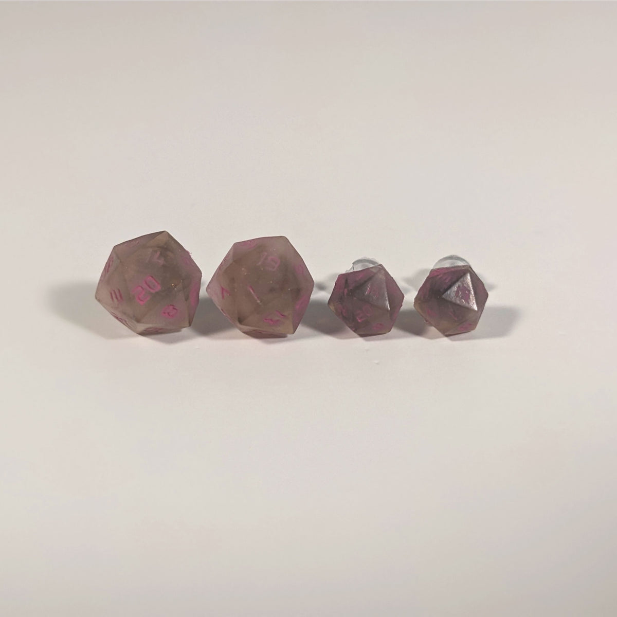 Two pairs of handmade d20 earrings with titanium hardware that are brown and white swirled with pink numbers on a white background sit in a line with the larger pair on the left and the smaller pair on the right. The earrings on the left of each pair centers the 20 face and the earrings on the right of each pair centers the 1 face.