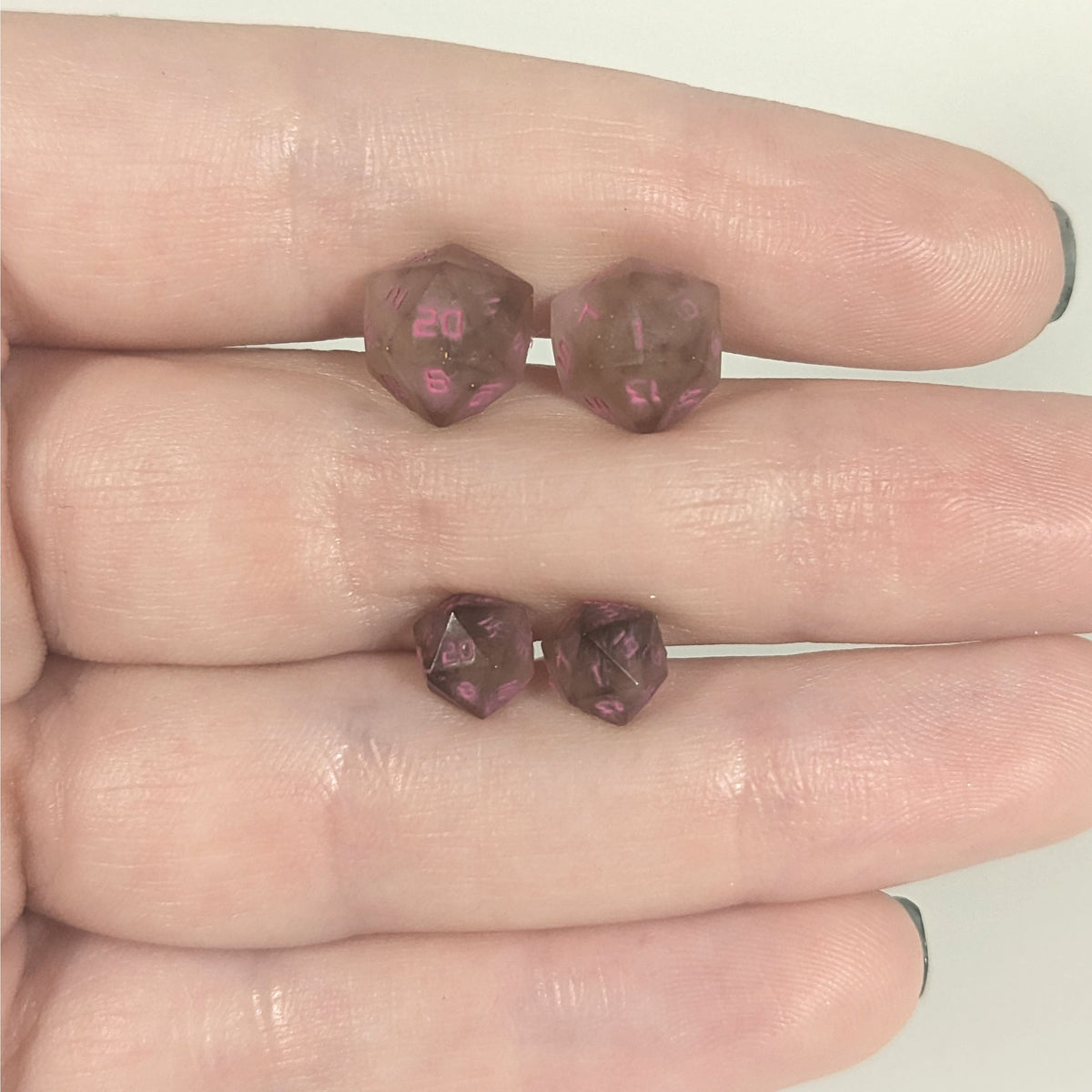 Two pairs of handmade d20 earrings with titanium hardware that are brown and white swirl with pink numbers held between the fingers of a flat hand. The larger pair is between the pointer and middle finger, the smaller between the middle and ring finger. The earrings on the left of each pair centers the 20 face and the earrings on the right of each pair centers the 1 face.