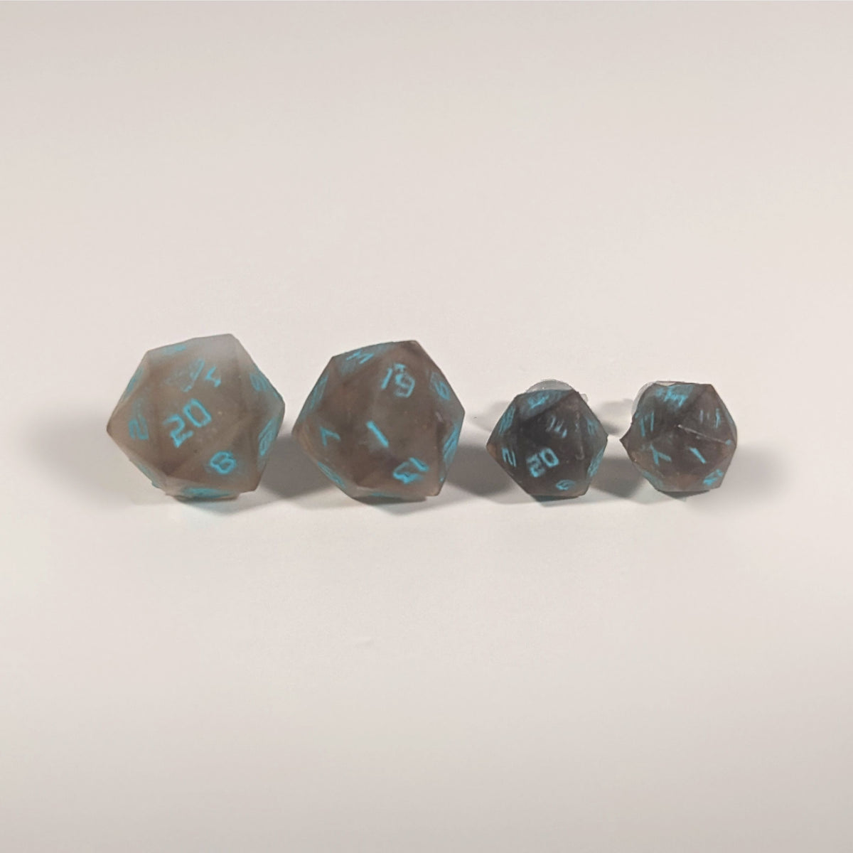 Two pairs of handmade d20 earrings with titanium hardware that are brown and white swirled with teal numbers on a white background sit in a line with the larger pair on the left and the smaller pair on the right. The earrings on the left of each pair centers the 20 face and the earrings on the right of each pair centers the 1 face.