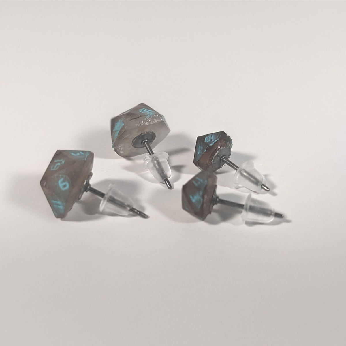 Two pairs of handmade d20 earrings that are brown and white swirled with teal numbers turned to show the titanium hardware and bell shaped rubber backs.