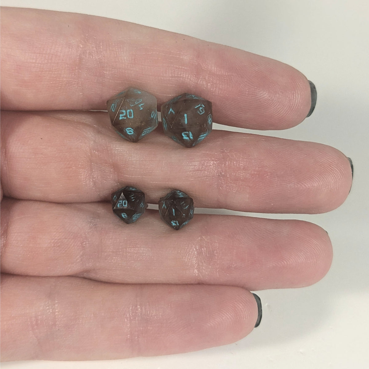 Two pairs of handmade d20 earrings with titanium hardware that are brown and white swirl with teal numbers held between the fingers of a flat hand. The larger pair is between the pointer and middle finger, the smaller between the middle and ring finger. The earrings on the left of each pair centers the 20 face and the earrings on the right of each pair centers the 1 face.