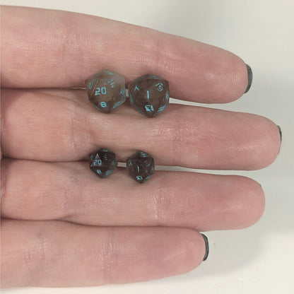 Two pairs of handmade d20 earrings with titanium hardware that are brown and white swirl with teal numbers held between the fingers of a flat hand. The larger pair is between the pointer and middle finger, the smaller between the middle and ring finger. The earrings on the left of each pair centers the 20 face and the earrings on the right of each pair centers the 1 face.