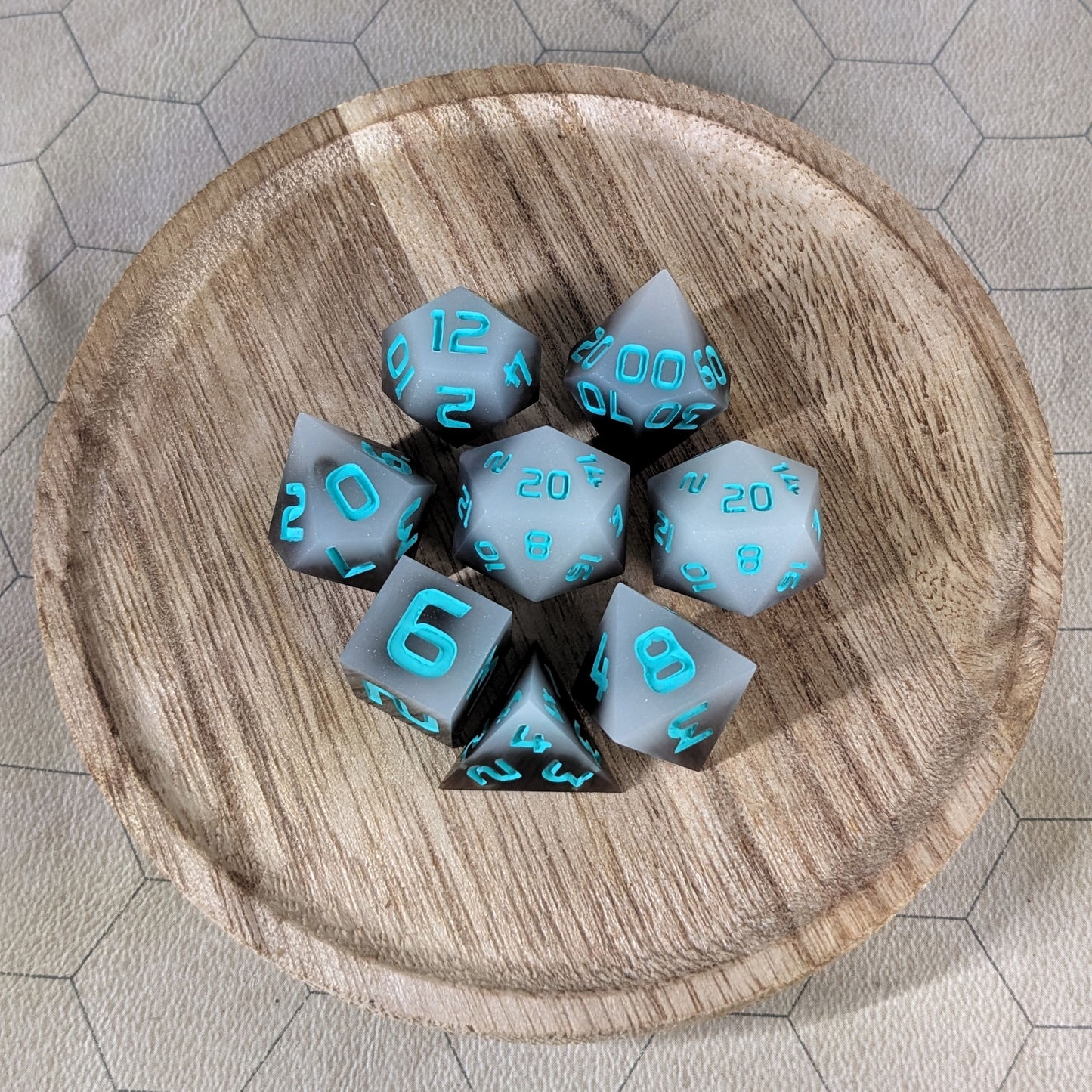 A complete set of 8 handmade artisan dice that are coffee brown with milky white swirls and teal numbers. The DnD dice set is on a wooded circle and the dice are arranged in a circle and features 2 D20s, one of which is in the center surrounded by the other 7 dice.