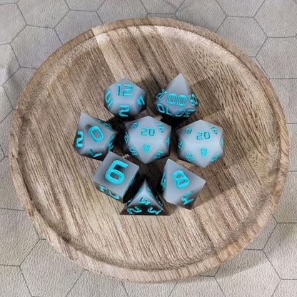 A complete set of 8 handmade artisan dice that are coffee brown with milky white swirls and teal numbers. The DnD dice set is on a wooded circle and the dice are arranged in a circle and features 2 D20s, one of which is in the center surrounded by the other 7 dice.