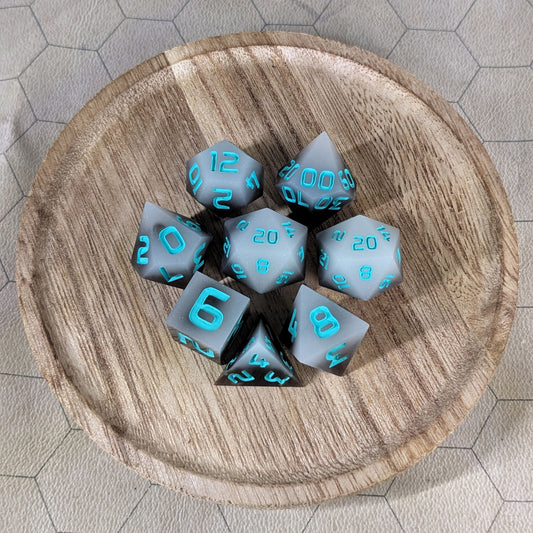 A complete set of 8 handmade artisan dice that are coffee brown with milky white swirls and teal numbers. The DnD dice set is on a wooded circle and the dice are arranged in a circle and features 2 D20s, one of which is in the center surrounded by the other 7 dice.