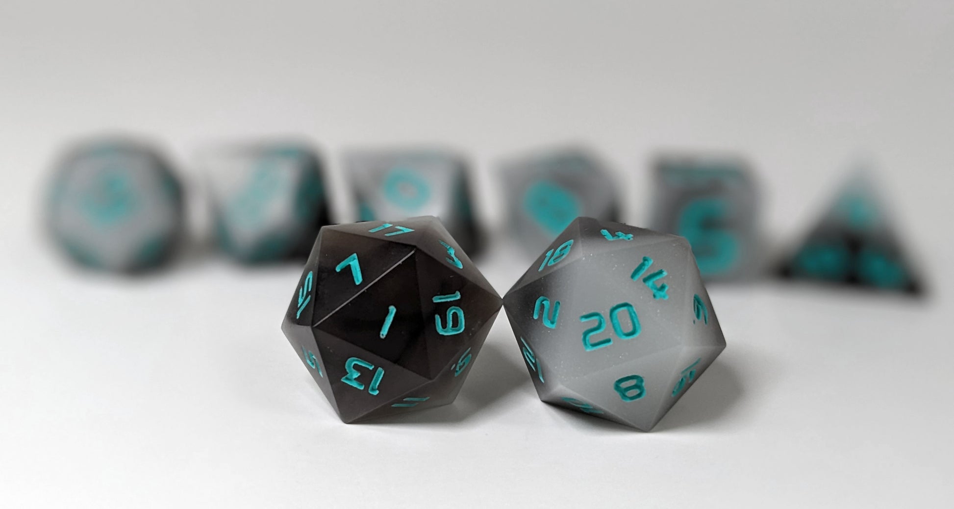 A straight on photo of a standard set of DnD dice that are coffee brown with milky white swirls and teal numbers focusing on 2 d20s in the center. The d20 on the left has the 1 face centered and the d20 on the right has the 20 face centered. The other 6 dice are lined up in the background but are out of focus, while the d20s are in sharp focus.