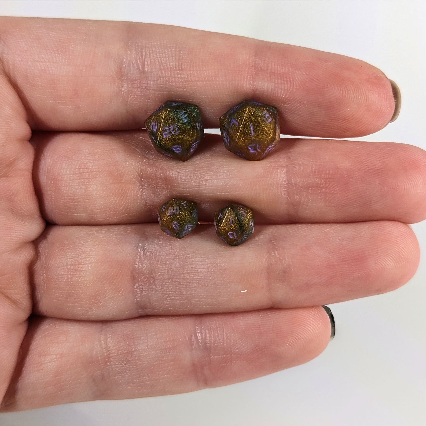 Two pairs of handmade d20 earrings with sterling silver hardware that are dark green and red gold with purple numbers held between the fingers of a flat hand. The larger pair is between the pointer and middle finger, the smaller between the middle and ring finger. The earrings on the left of each pair centers the 20 face and the earrings on the right of each pair centers the 1 face.