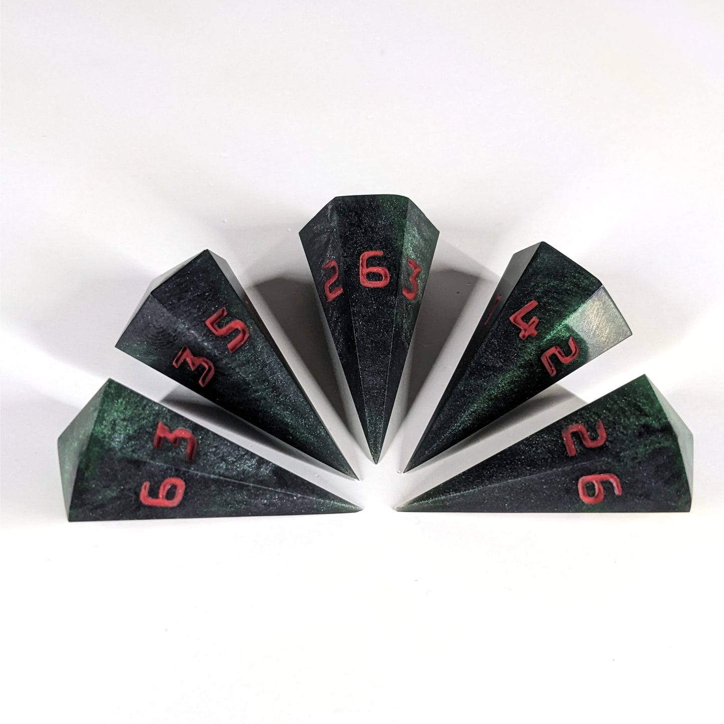 Five dagger shaped d6s are arranged in a semicircle with all the points of the dice pointing in towards a central point on a white background. The dice are black and dark green with red numbers, and the face up numbers from left to right (clockwise) are 3, 5, 6, 4, and 2.