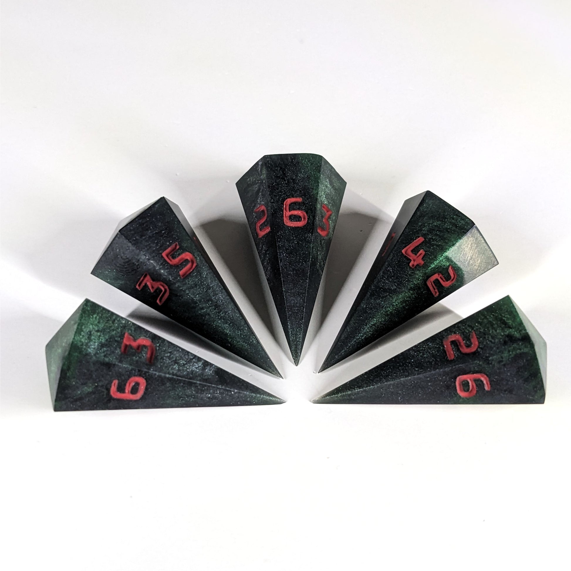 Five dagger shaped d6s are arranged in a semicircle with all the points of the dice pointing in towards a central point on a white background. The dice are black and dark green with red numbers, and the face up numbers from left to right (clockwise) are 3, 5, 6, 4, and 2.