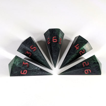 Five dagger shaped d6s are arranged in a semicircle with all the points of the dice pointing in towards a central point on a white background. The dice are black and dark green with red numbers, and the face up numbers from left to right (clockwise) are 3, 5, 6, 4, and 2.