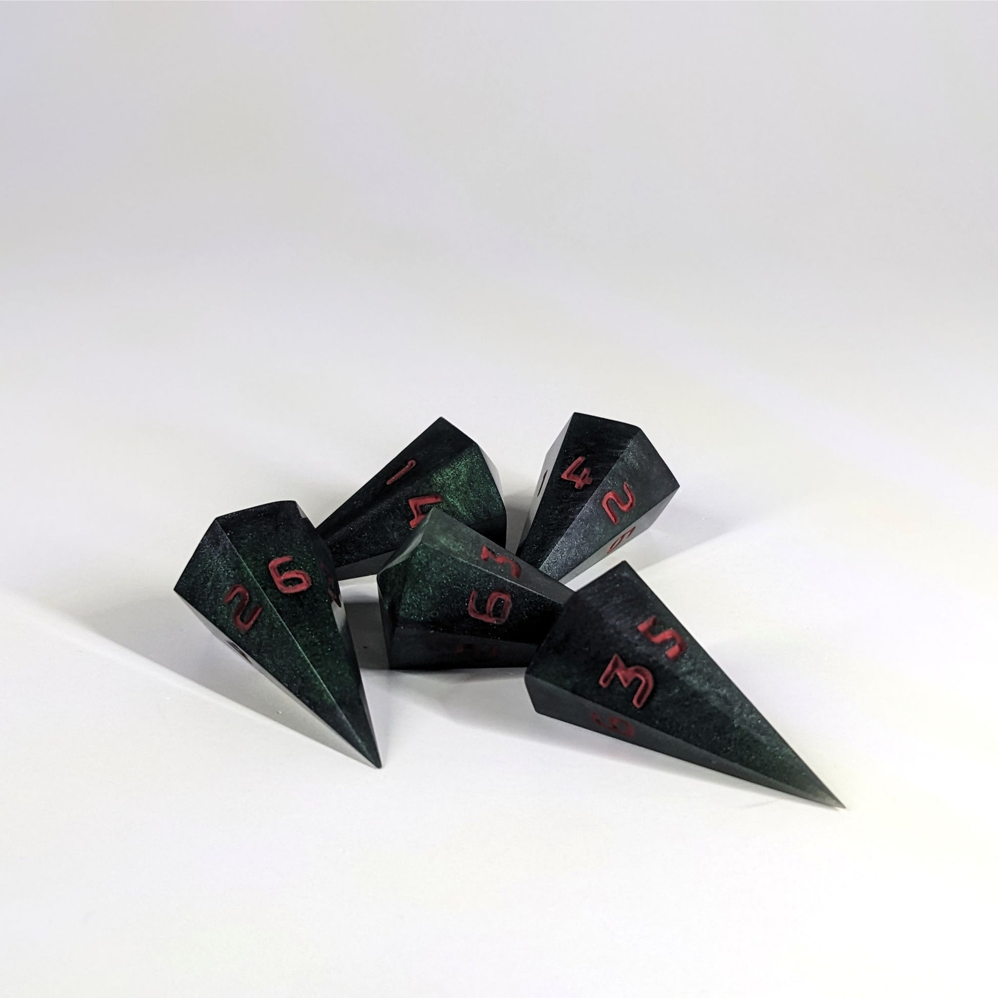 Five dagger shaped d6s are scattered on a white background. The handmade dice are black and dark green with red numbers.