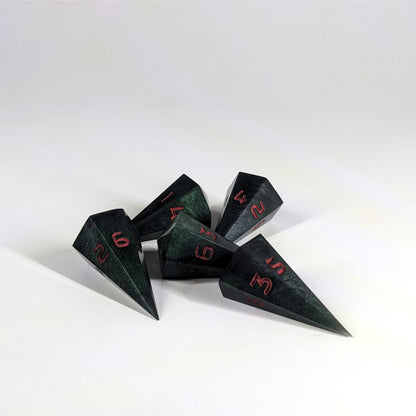 Five dagger shaped d6s are scattered on a white background. The handmade dice are black and dark green with red numbers.