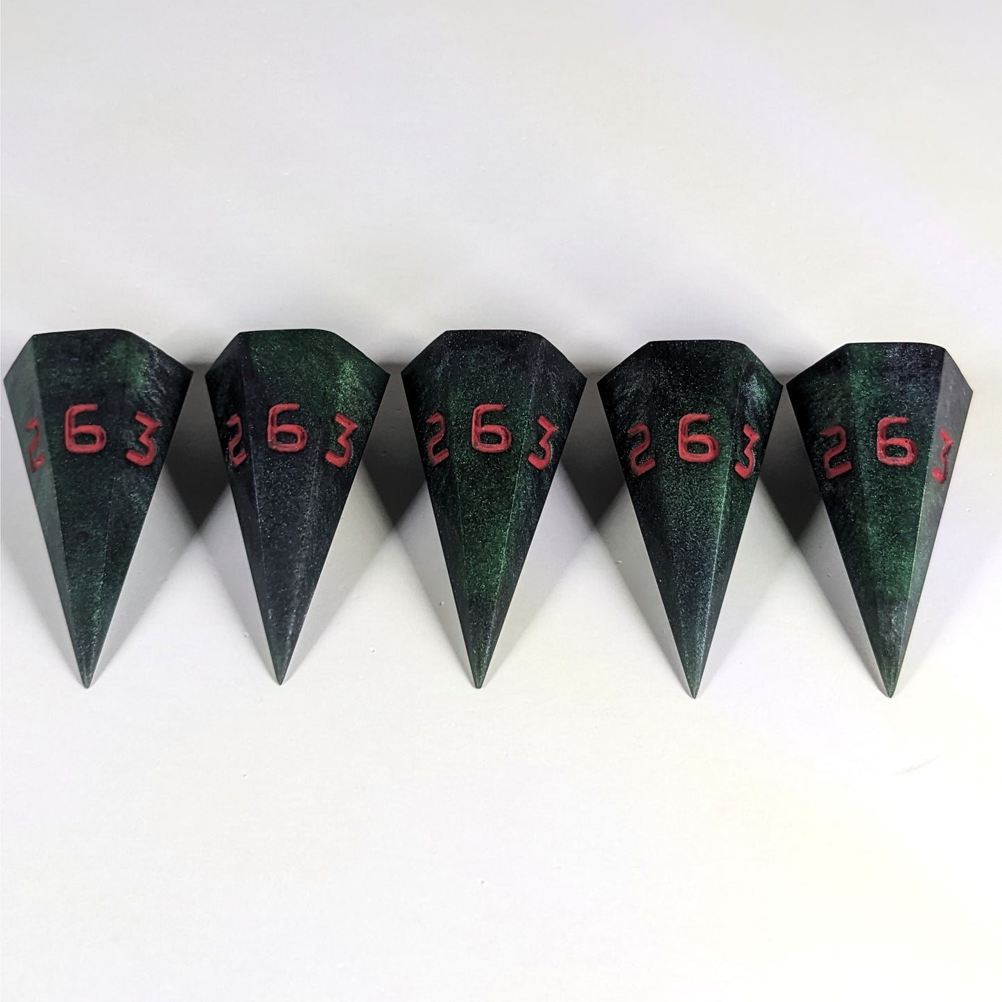 Five dagger shaped d6s are arranged in a line with the points of the dice facing downwards. The dice are dark green and black with red numbers, and they all have the 6 face up.