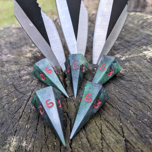 Five dagger shaped d6s sit on a stump with 3 throwing knives stabbed into the wood behind them. The dice are black and dark green with red numbers, and all have the 6 face up.