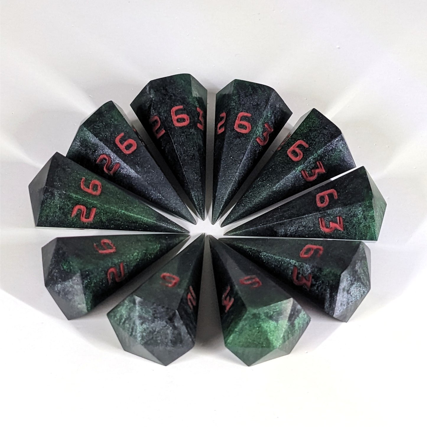 Ten dagger shaped d6s are arranged in a circle with all the points of the dice pointing in towards a central point on a white background. The dice are black and dark green with red numbers, and the face up numbers are all 6.