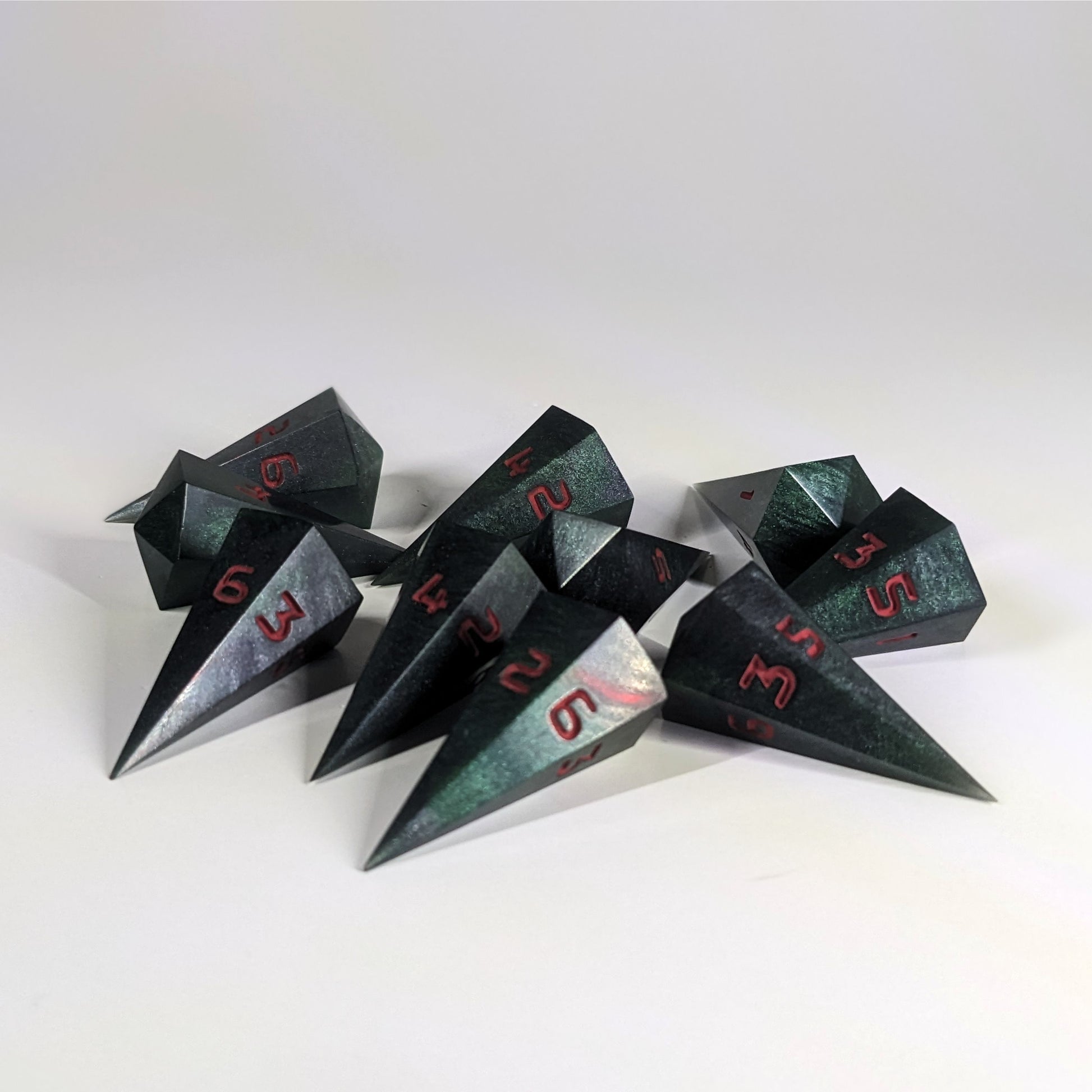 Ten dagger shaped d6s are scattered on a white background. The handmade dice are black and dark green with red numbers.