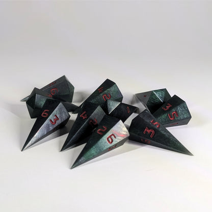 Ten dagger shaped d6s are scattered on a white background. The handmade dice are black and dark green with red numbers.