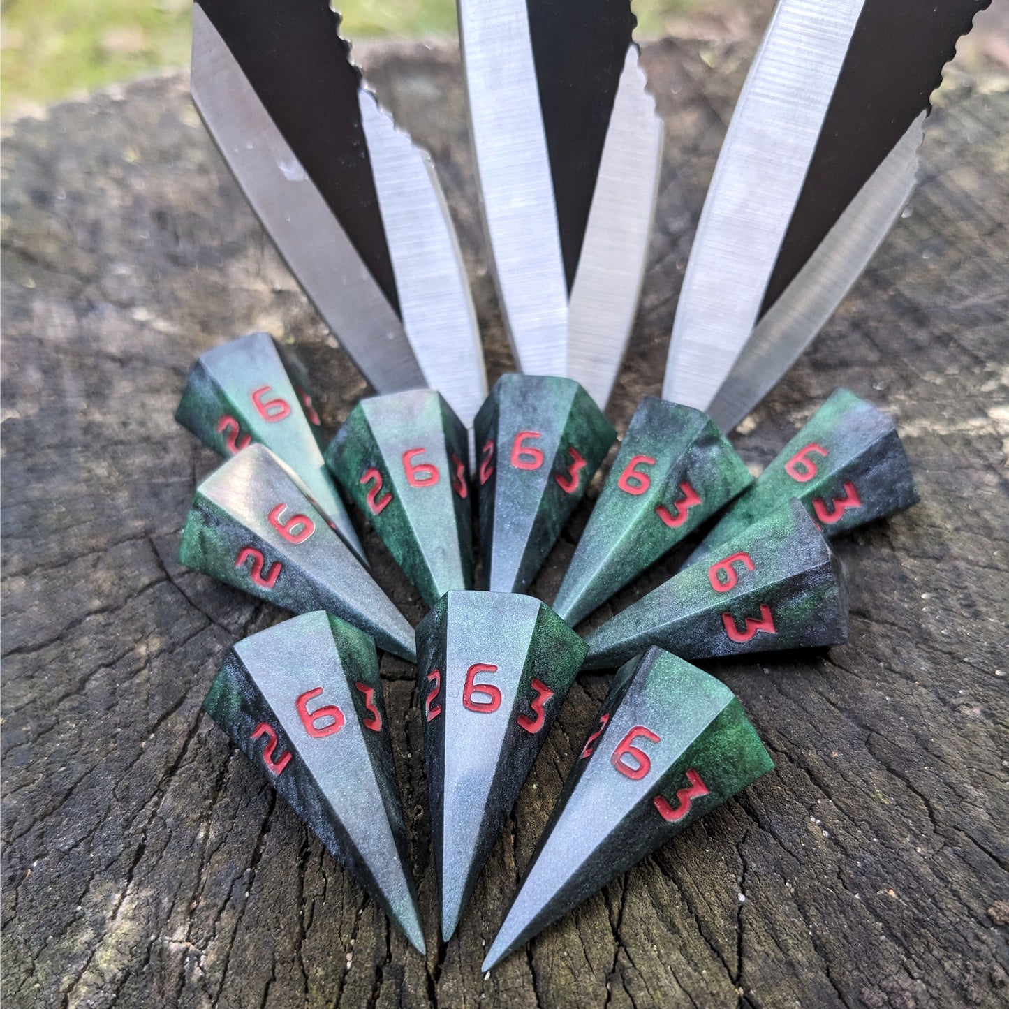 Ten dagger shaped d6s sit on a stump with 3 throwing knives stabbed into the wood behind them. The dice are black and dark green with red numbers, and all have the 6 face up.