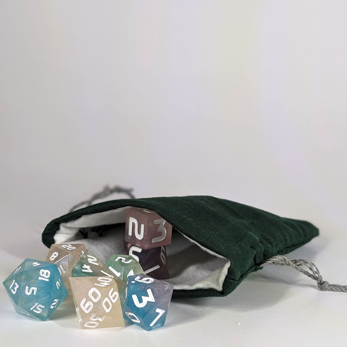 A small, handmade dice bag that is dark green lays open with a set of colorful, dnd dice with white numbers spilling out of it on a white background.