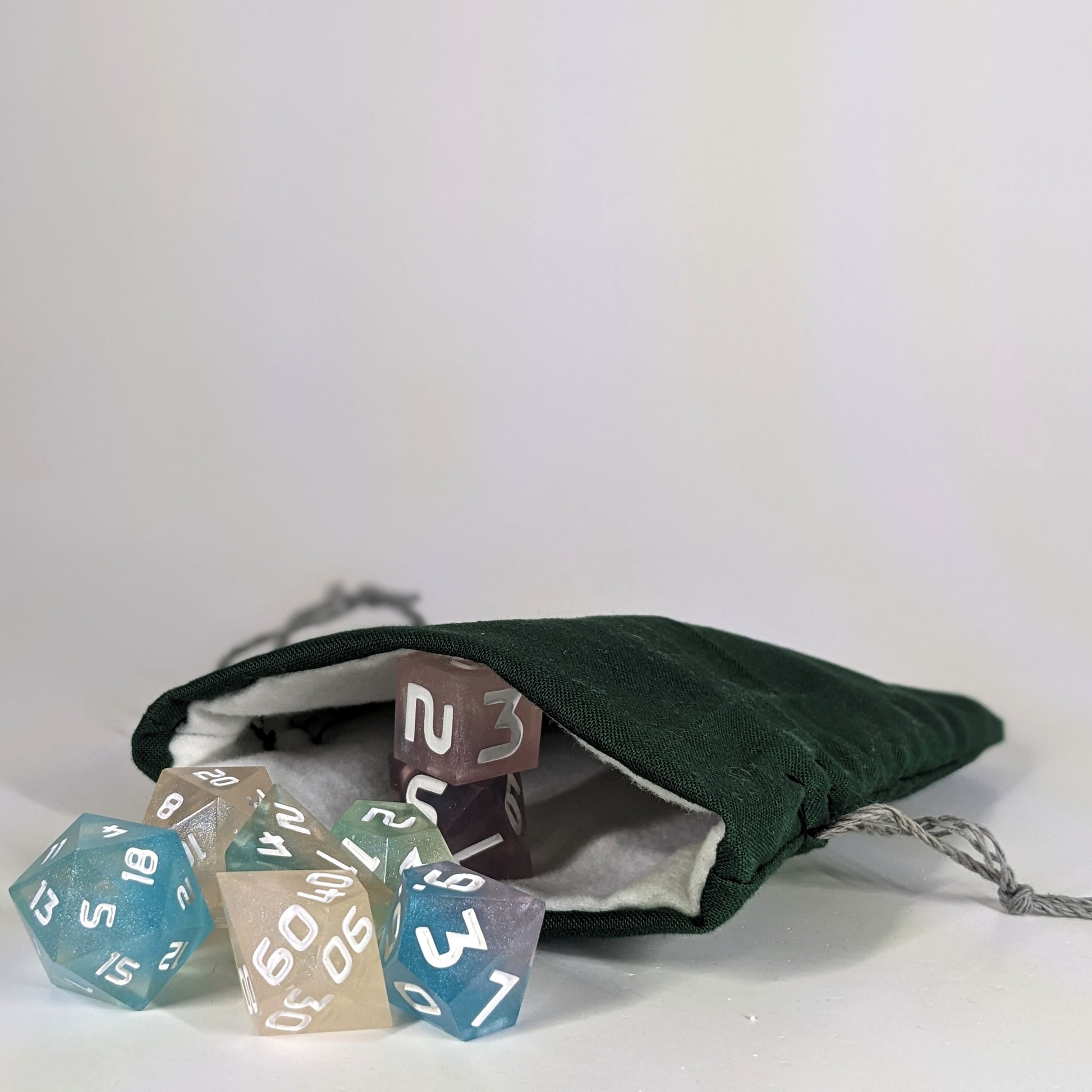 A small, handmade dice bag that is dark green lays open with a set of colorful, dnd dice with white numbers spilling out of it on a white background.