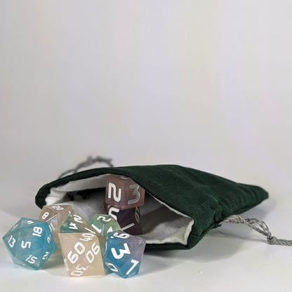 A small, handmade dice bag that is dark green lays open with a set of colorful, dnd dice with white numbers spilling out of it on a white background.