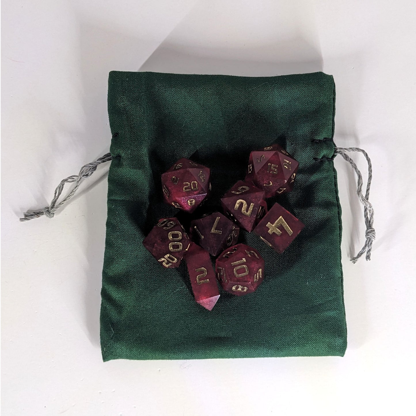 A small, handmade dice bag that is dark green lays flat on a white background with a set of red dnd dice with bronze numbers sitting on top of it.