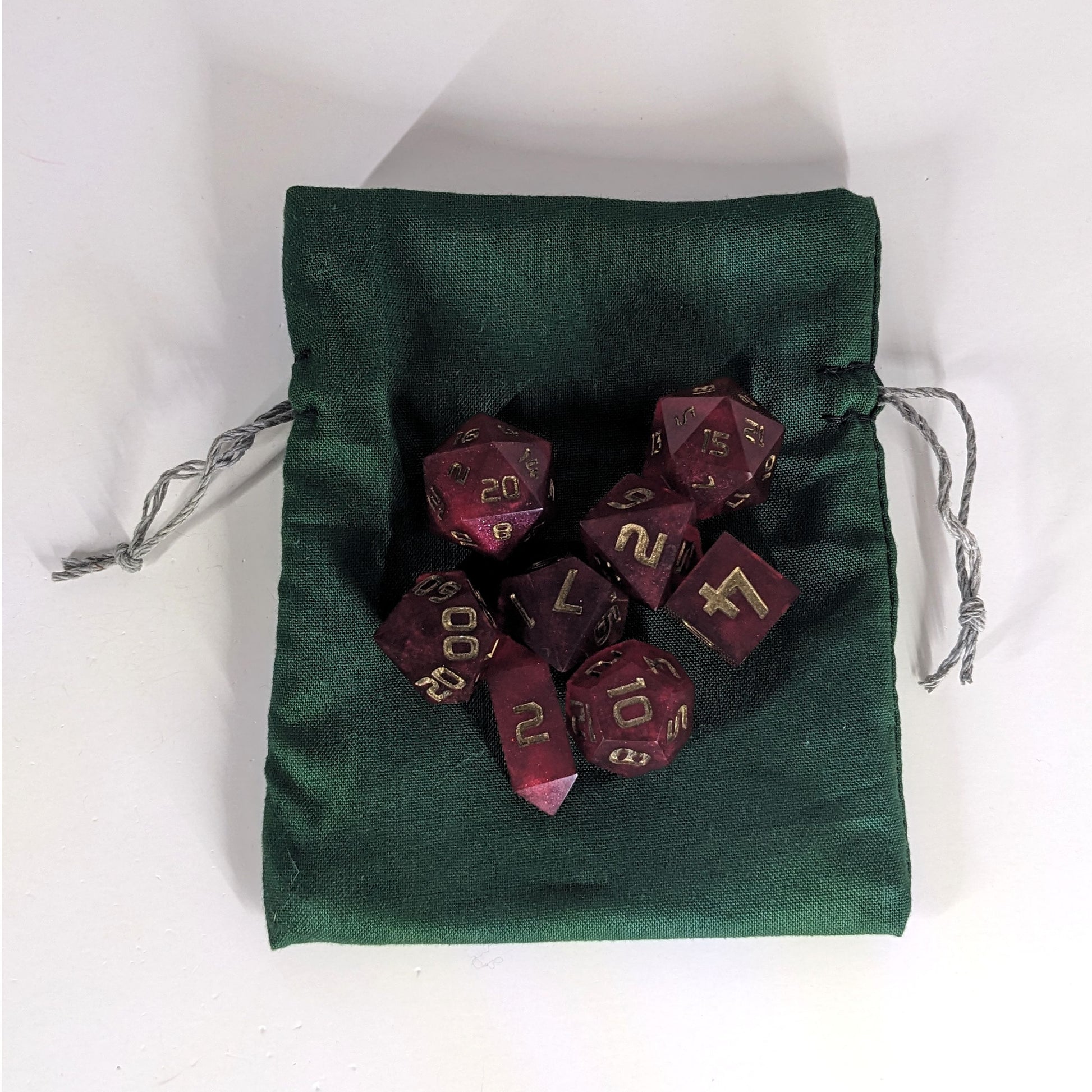 A small, handmade dice bag that is dark green lays flat on a white background with a set of red dnd dice with bronze numbers sitting on top of it.