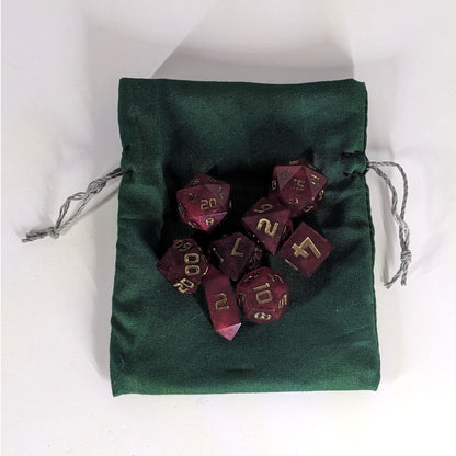 A small, handmade dice bag that is dark green lays flat on a white background with a set of red dnd dice with bronze numbers sitting on top of it.